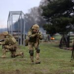 Група військових рухається вперед по відкритій місцевості, відпрацьовуючи бойову взаємодію. Один із них прикриває просування, тримаючи зброю напоготові.