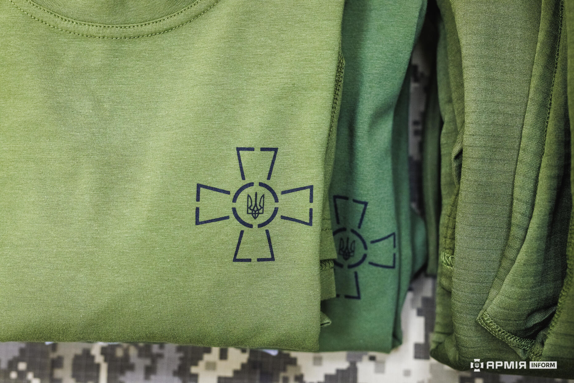 Складені зелені футболки з нанесеним символом у вигляді хреста з тризубом у центрі лежать одна на одній. Акцент зроблено на чіткості принта та однорідності тканини.