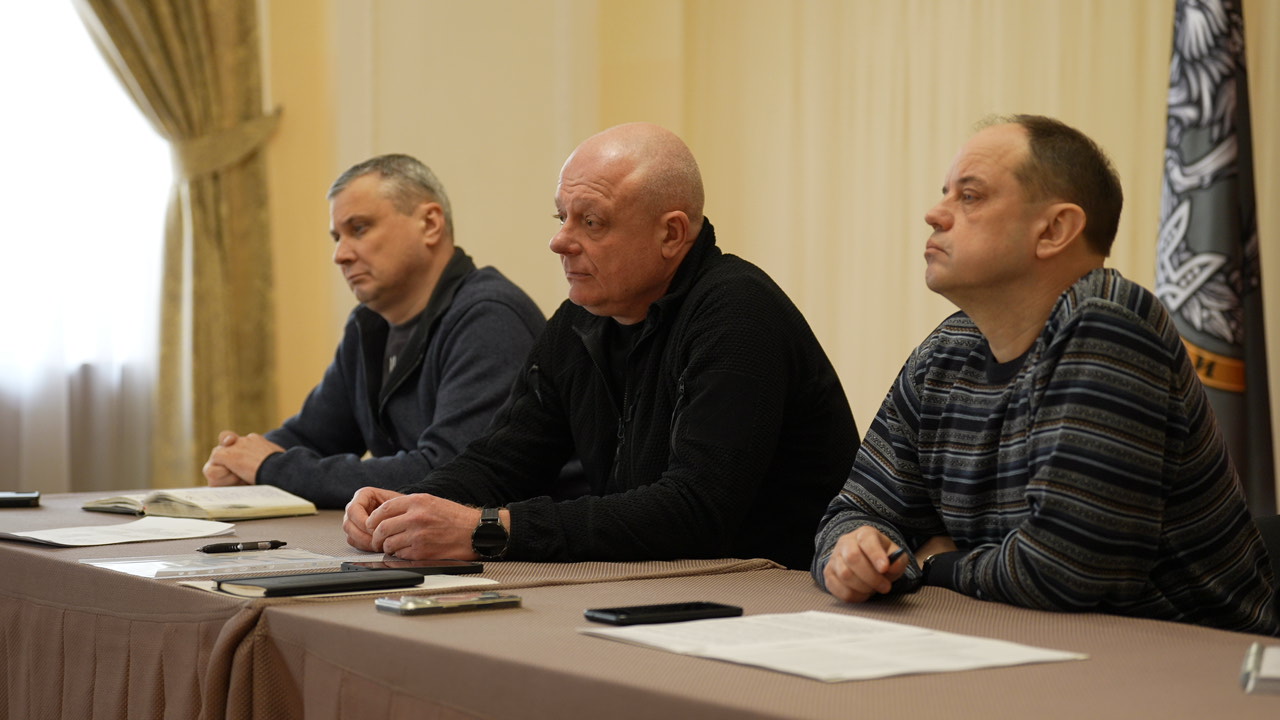 Троє чоловіків за столом уважно слухають і готуються до обговорення, перед ними — записи та телефони. Вони виконують роль спікерів або організаторів заходу.