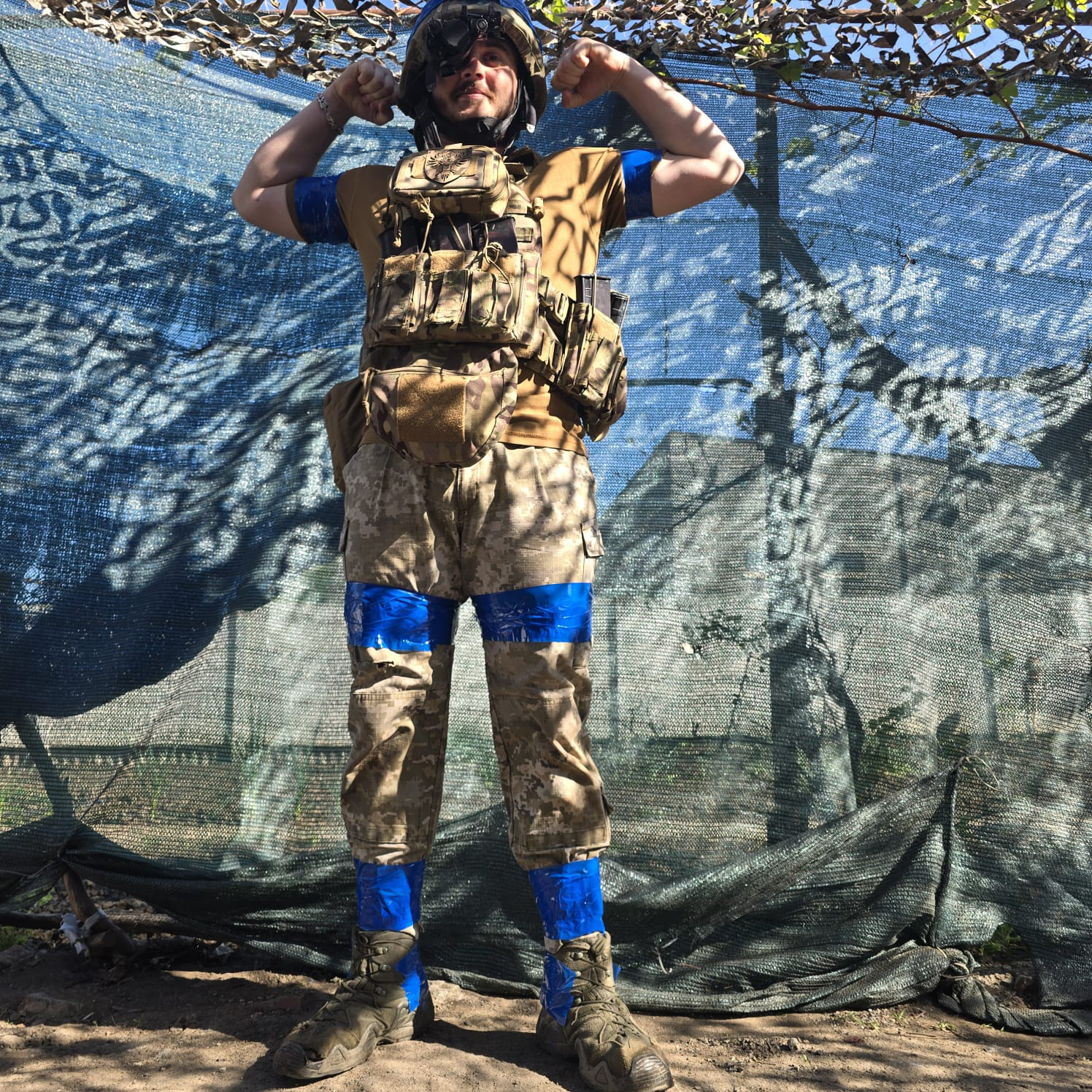 Військовий у бронежилеті та касці стоїть під маскувальною сіткою й демонструє м’язи, піднявши обидві руки. На ногах і руках видно синю стрічку, а навколо — польові укриття.