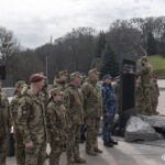 Військовий, стоячи на кам’яній платформі, додає новий шеврон до загальної композиції. Інші учасники церемонії стоять поруч у строю, спостерігаючи за дією.