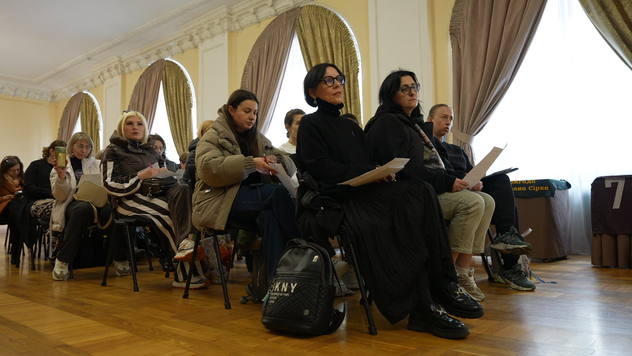 Жінки в залі слухають виступ і тримають у руках роздаткові матеріали. Атмосфера робоча, з акцентом на навчання та обговорення.