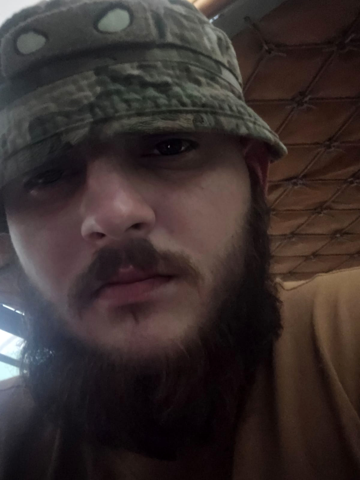 Крупний план обличчя чоловіка у панамі, який дивиться прямо в камеру. Освітлення слабке, а фон нагадує внутрішнє приміщення з тканинним або сітчастим покриттям.