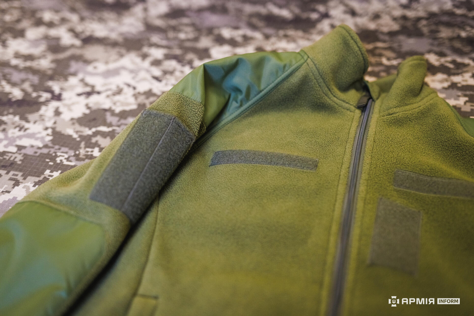 Зелена тактична кофта з блискавкою показана крупним планом із панелями на липучках для шевронів. Матеріал виглядає щільним і призначеним для використання в польових умовах.