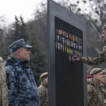 Ще один військовослужбовець прикріплює шеврон до стенда, зосереджено виконуючи дію. Поруч стоять колеги у формі, серед них видно бійців у беретах різних кольорів.