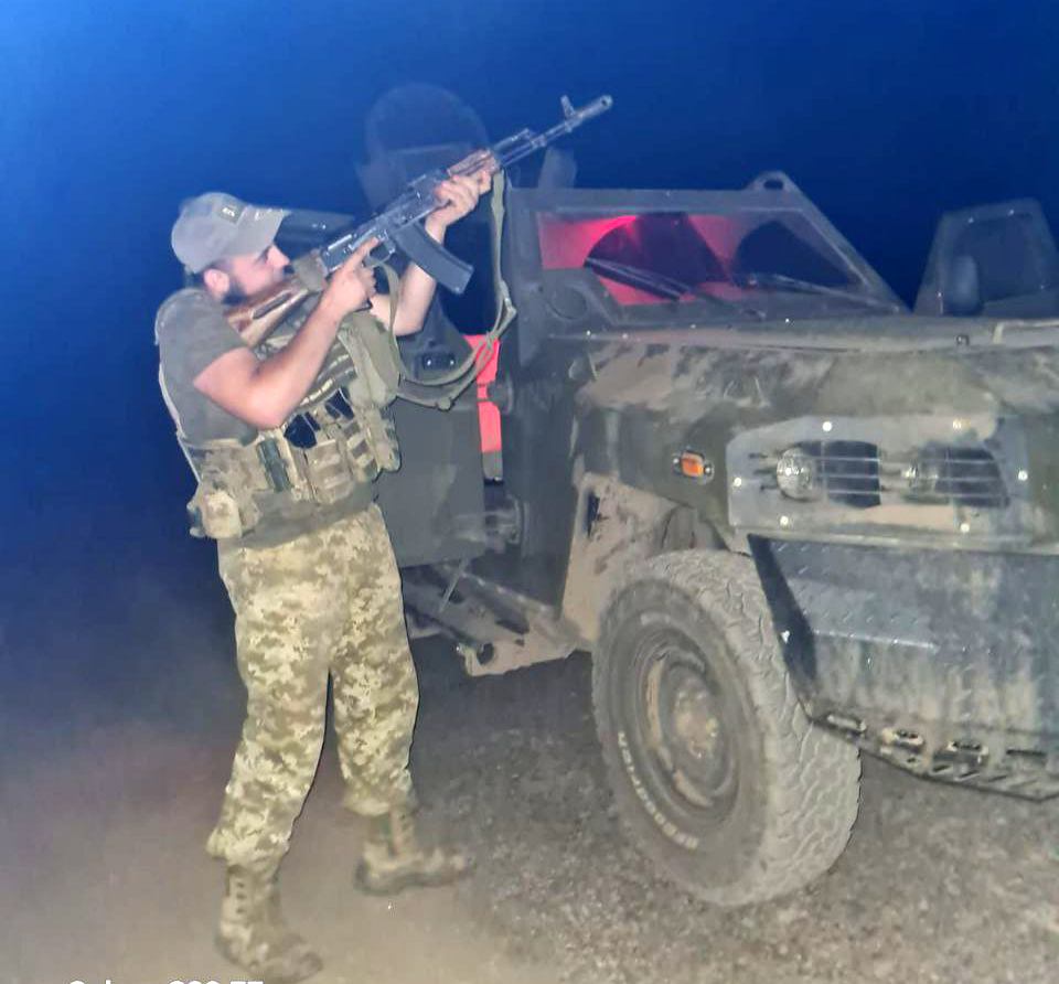 Військовий стоїть поруч із броньованим автомобілем уночі та тримає автомат, піднятий угору. Машина має пошкоджений або зношений вигляд, а навколо темрява і світло фар.