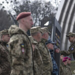 Військовий у формі з окулярами додає шеврон до стенда, нахилившись ближче для точності. Інші військові стоять поруч, утворюючи лінію учасників церемонії.