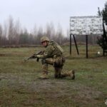 Боєць на коліні тримає автомат напоготові, уважно стежачи за умовною ціллю попереду. Позаду видно навчальну інфраструктуру та інформаційний стенд.