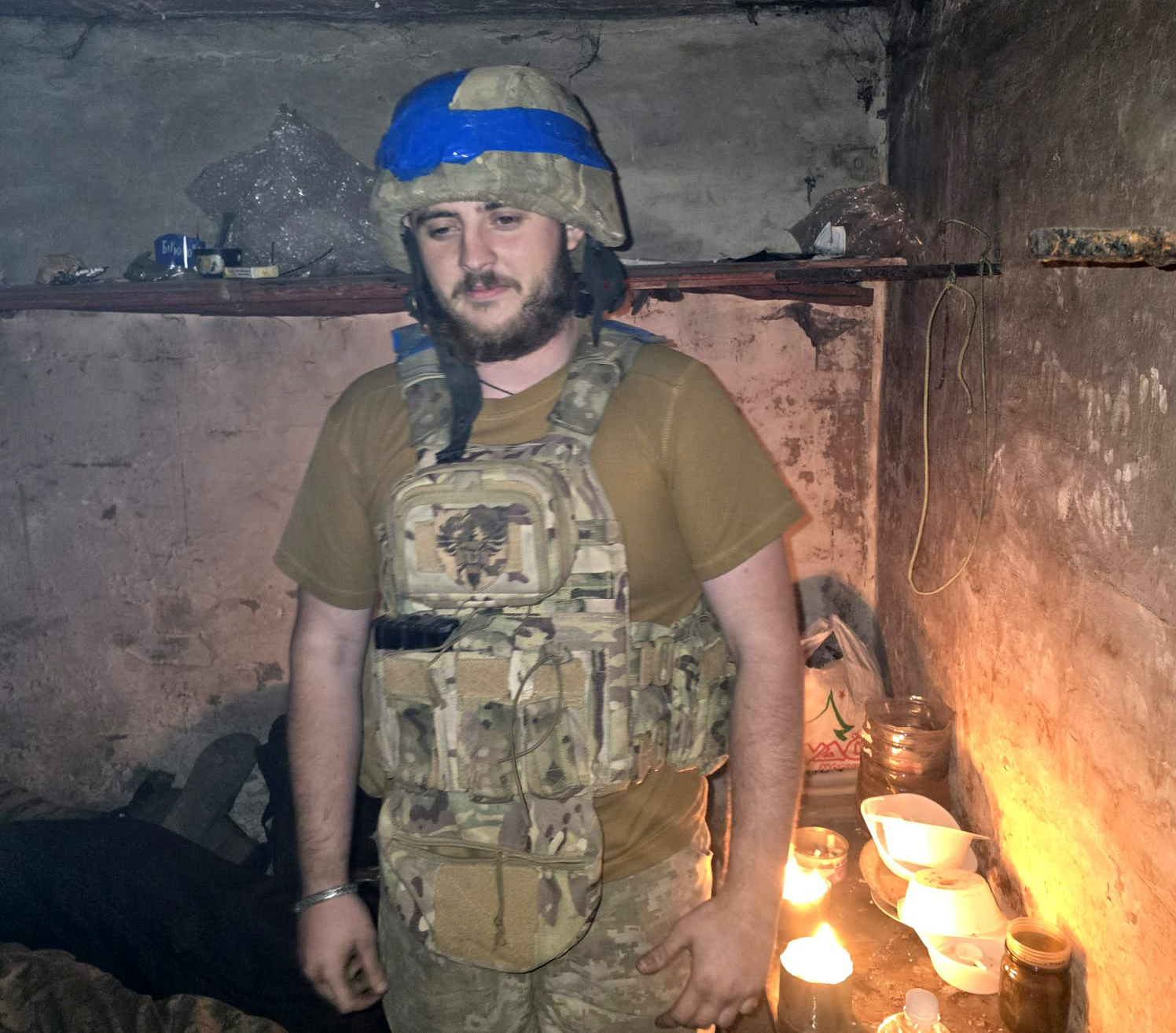 Боєць у бронежилеті перебуває в приміщенні з грубими стінами, де горять свічки та стоїть посуд. Атмосфера нагадує імпровізоване укриття або бліндаж.