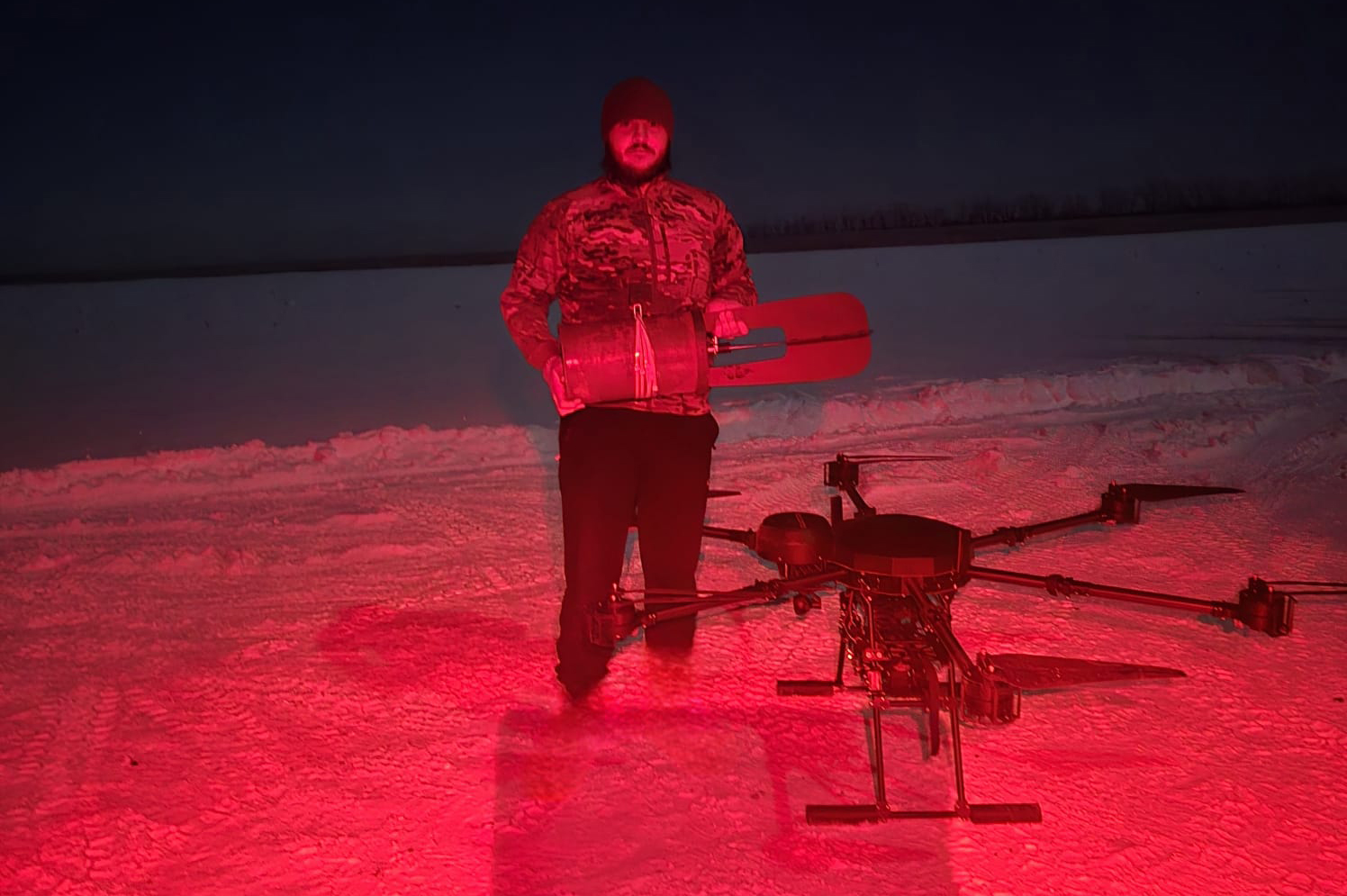 Чоловік стоїть на снігу поруч із великим дроном, освітленим червоним світлом. Він тримає в руках обладнання, а навколишній простір виглядає відкритим і нічним.