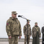 Військовий виступає перед мікрофоном на відкритому майданчику, за ним стоять представники духовенства та інші учасники. У повітрі відчувається офіційна й стримана атмосфера заходу.
