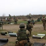 Група військових у захисному спорядженні працює на стрільбищі, ведучи вогонь по мішенях. Інструктори спостерігають і коригують дії бійців.