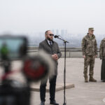 Цивільний у пальті виступає з промовою перед мікрофоном, на фоні — військові та священник. У кадрі також видно камеру, що знімає подію.