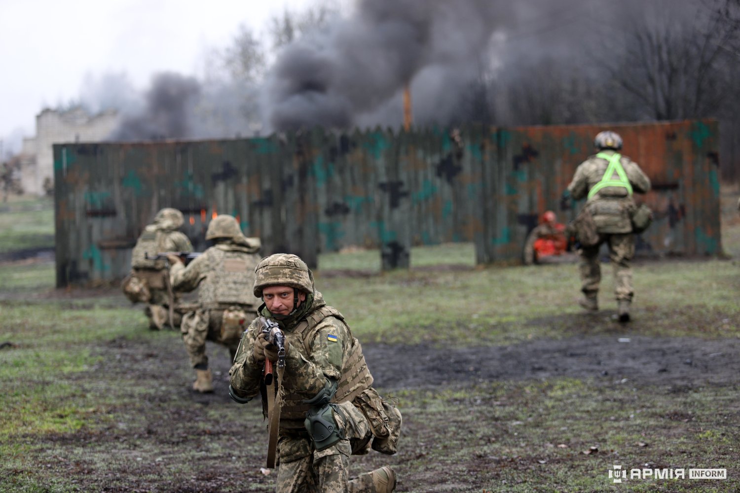 Боєць у передньому плані рухається вперед із автоматом, прикриваючи товаришів, які наступають позаду. У фоні видно дим і умовні осередки вогню, що імітують бойову ситуацію.