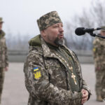 Священнослужитель у військовій формі з хрестом на грудях говорить у мікрофон. Його виступ відбувається на тлі військових, які стоять позаду в строю.