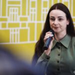 Жінка виступає перед аудиторією з мікрофоном на тлі виставкового або конференц-простору. Імовірно, вона проводить презентацію чи екскурсію, присвячену представленій історичній експозиції.
