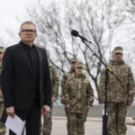 Чоловік у чорному костюмі стоїть біля мікрофона, тримаючи аркуш паперу, позаду — військові у строю. Його поза й вираз обличчя підкреслюють офіційний і стриманий характер виступу.