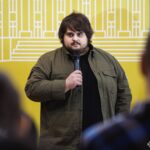 На фото молодий чоловік виступає перед аудиторією з мікрофоном на тлі стилізованої виставкової стіни. Захід має формат публічної презентації або церемонії, присвяченої історичній тематиці.