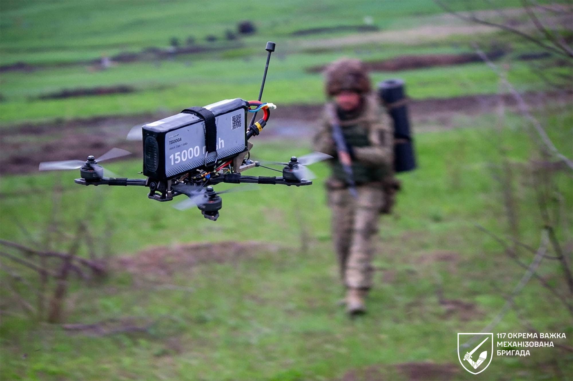 У кадрі — FPV-дрон із потужним акумулятором, що завис у повітрі на передньому плані. На розмитому тлі військовослужбовець у спорядженні рухається полем, готуючись до виконання завдання.