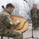 Військовослужбовець сидить і грає на бандурі, зосереджено перебираючи струни. На задньому плані стоять інші військові, слухаючи музичний виступ.