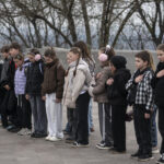 Група дітей стоїть у ряд із руками на грудях, дотримуючись урочистого моменту. Їхні серйозні обличчя свідчать про повагу до події.