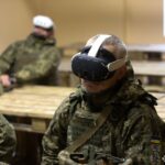 Кілька військових сидять у приміщенні, використовуючи VR-гарнітури під час навчання. Обстановка класу й лави підкреслюють організований тренувальний процес.