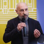 Чоловік у діловому одязі виступає перед гостями, акцентуючи увагу на темі заходу. Формальна атмосфера підкреслює значущість презентації або виставки.