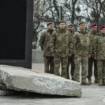 Військові стоять у строю поруч із меморіальним стендом і уламками бетонних плит. Атмосфера зосереджена й урочиста, всі дивляться вперед.