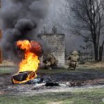 На передньому плані палає імпровізована перешкода, піднімаючи густий чорний дим. Двоє військових за укриттям відпрацьовують дії в умовах наближених до бою.