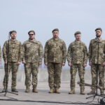 Група військових у беретах співає перед мікрофонами, виконуючи, ймовірно, патріотичну композицію. Їхні обличчя напружені, а постава — зібрана й синхронна.