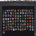 Стенд із великою кількістю шевронів різних підрозділів заповнює центр кадру. Кожен знак відображає окрему частину або підрозділ, утворюючи єдину композицію.