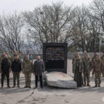 Учасники заходу вишикувалися по обидва боки від стенда з шевронами та уламками плит. Серед них — військові, цивільні та священнослужитель, що створює спільну урочисту сцену.