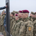 Військові в беретах стоять у строю, спостерігаючи, як інший учасник прикріплює елемент до стенда. Їхні обличчя зосереджені, а постава — дисциплінована.