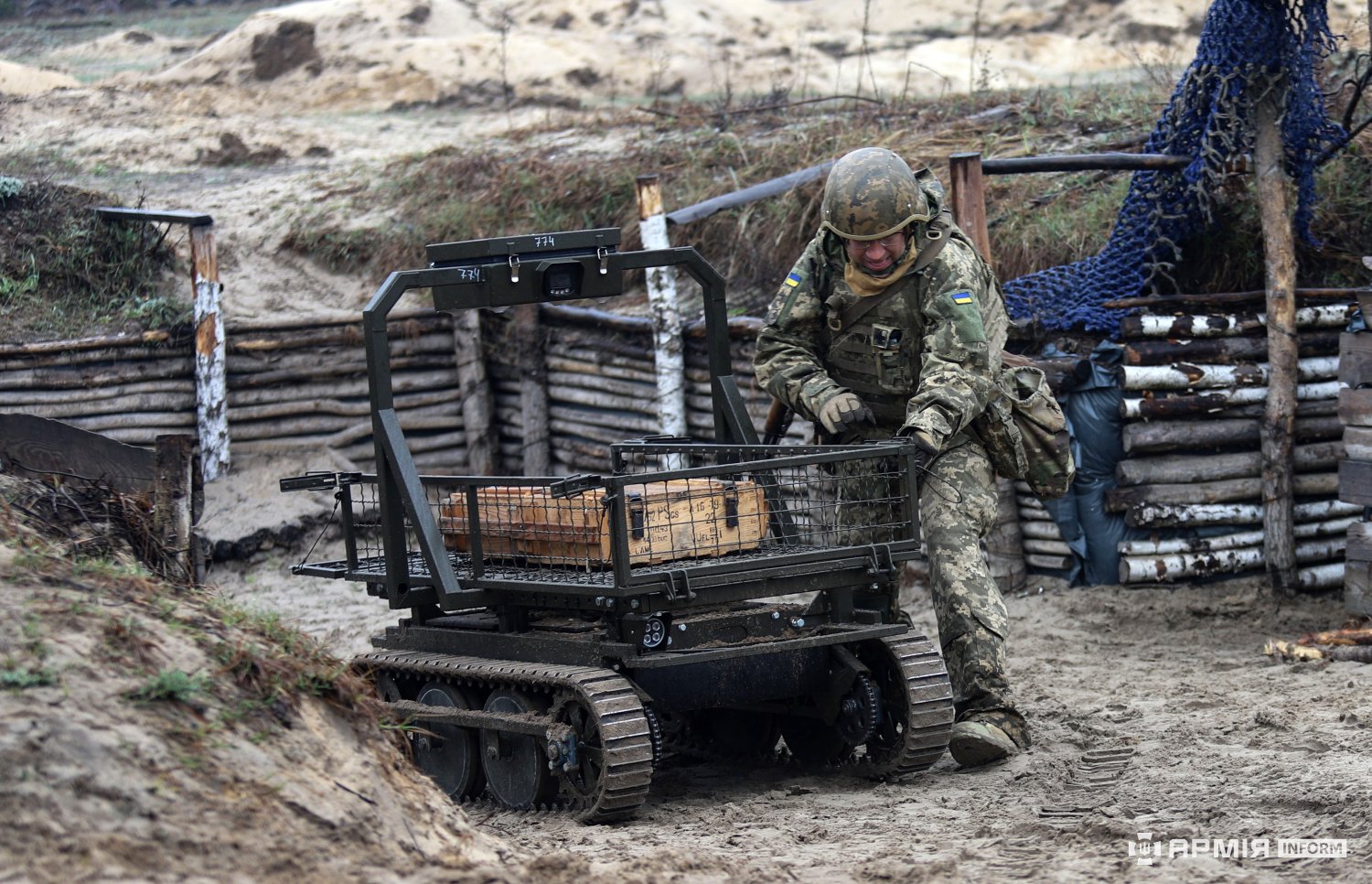 Боєць у траншеї керує гусеничною роботизованою платформою, завантаженою ящиками. Конструкція окопів і піщаний ґрунт підкреслюють польові умови використання техніки.