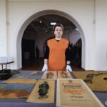 Жінка в рукавичках стоїть за експозиційним столом із колекцією історичних артефактів УНР. Вона, ймовірно, представляє або курує виставку, присвячену українському військовому спадку.