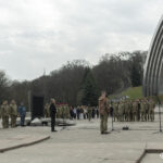 Загальний план показує місце проведення церемонії з великою кількістю військових, глядачів і журналістів. У центрі — стенд і виступаючий біля мікрофона, що об’єднує всіх присутніх.