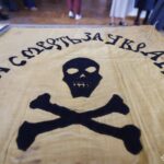 На знімку представлено старовинне бойове знамено з написом «На смерть за Україну» та символікою черепа з кістками. Прапор є яскравим прикладом військової символіки українських формувань початку ХХ століття.
