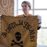 Жінка у захисних рукавичках демонструє історичне знамено з написом «На смерть за Україну». Артефакт подано як один із центральних експонатів виставки.