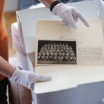 На фото кураторка в білих рукавичках демонструє архівну папку з історичними світлинами та документами. Серед матеріалів видно групове фото військового колективу, що підкреслює цінність збережених архівів для дослідження української історії.