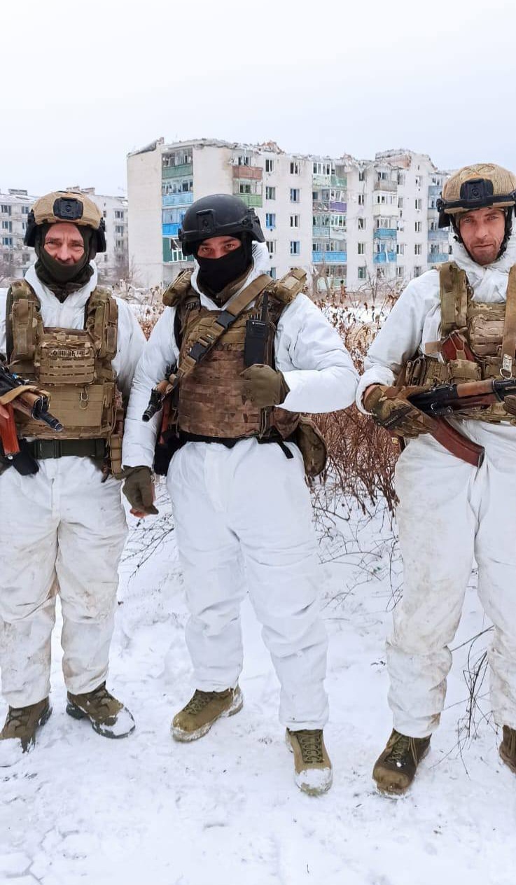 Троє українських військових у зимовому камуфляжі позують на фоні багатоповерхівок