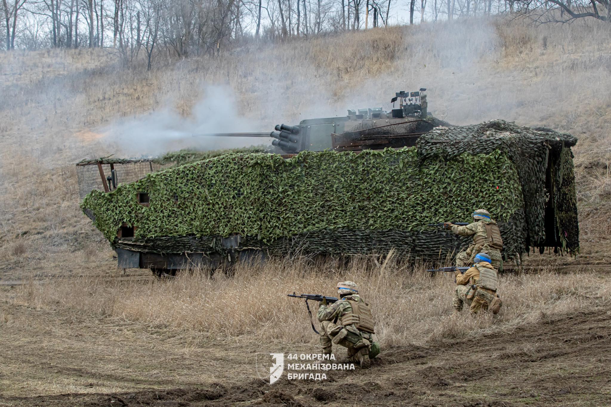 На позиції серед сухої трави замаскована бронемашина веде вогонь, здіймаючи дим із гармати. Попереду неї троє військових у захисному спорядженні займають позиції та прикривають техніку, готуючись до подальших дій.