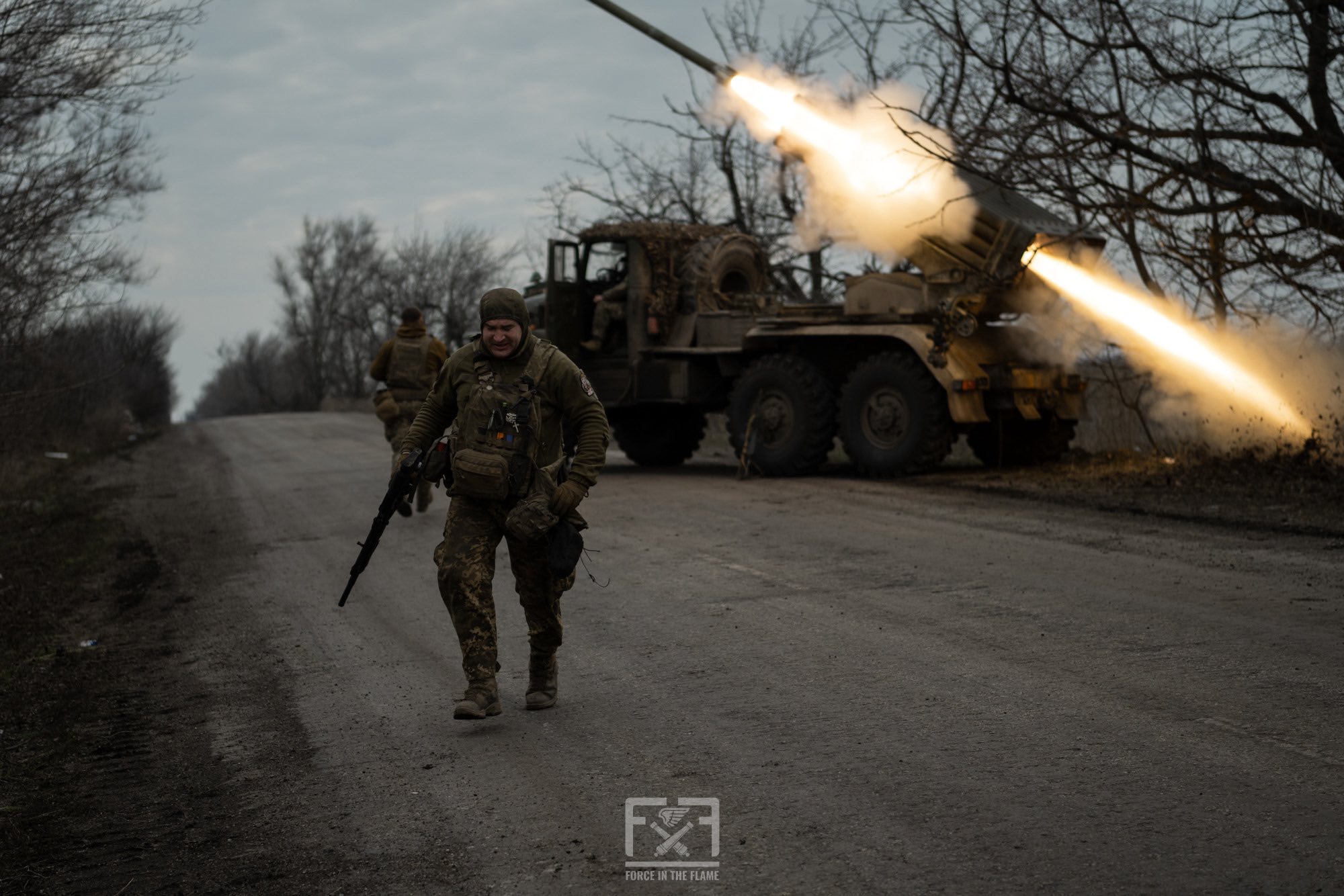 На дорозі військовий із автоматом швидко відходить від реактивної системи залпового вогню, яка саме здійснює пуск ракети. У сутінках видно спалах і дим від пострілу, а навколо — оголені дерева та ще один боєць біля техніки.