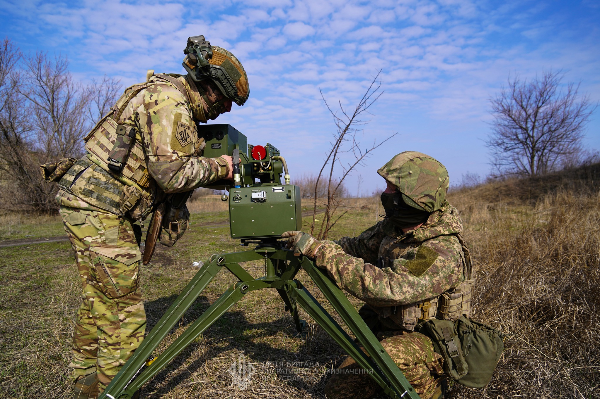 Двоє військових у польових умовах налаштовують протитанковий комплекс, встановлений на тринозі. Один працює з прицільним блоком, інший фіксує елементи установки, перебуваючи поруч на відкритій місцевості.