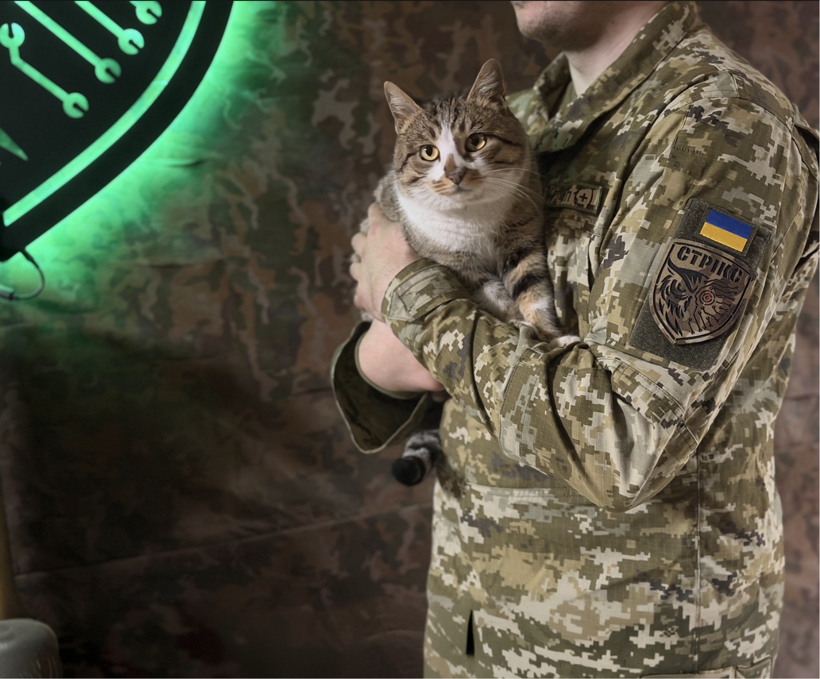 На фото військовослужбовець у камуфляжі тримає на руках спокійного смугастого кота, який уважно дивиться в об’єктив. На його рукаві чітко видно шеврон із прапором України та написом «СТРІКС», під яким зображено стилізовану голову сови з прицілом — символ пильності й нічного бачення. Тло з камуфльованою тканиною та неоновим знаком із технічними елементами підсилює атмосферу підрозділу, пов’язаного з технологіями або розвідкою.