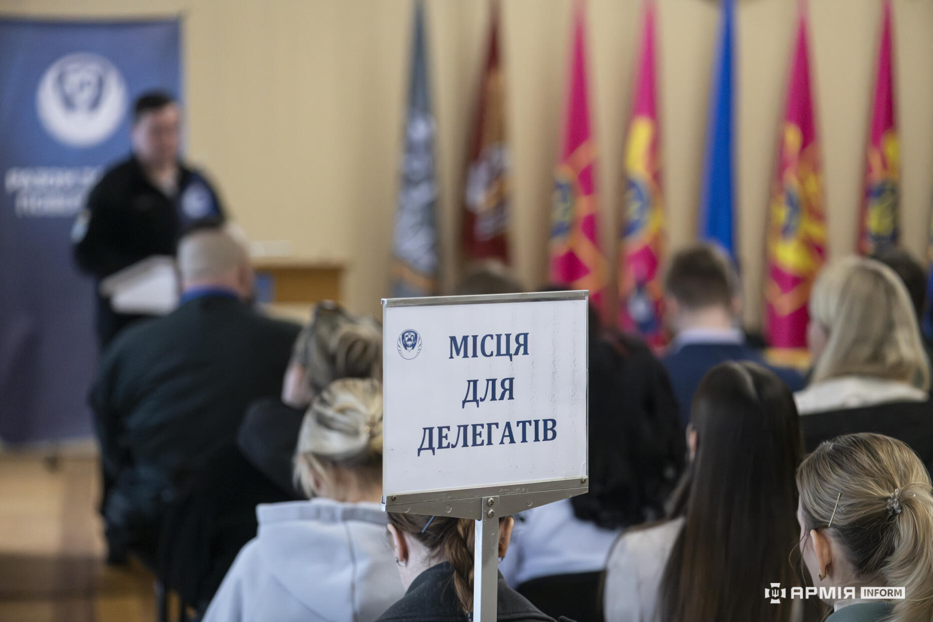 У залі під час офіційного заходу на передньому плані розміщено табличку «Місця для делегатів», тоді як доповідач виступає перед аудиторією. На тлі видно прапори, банер організації та учасників, зосереджених на перебігу події.