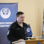 Чоловік у форменому одязі виступає за трибуною під час конференції або зборів, звертаючись до присутніх. Позаду нього розташований банер із написом «Разом заради повернення» та символіка заходу.