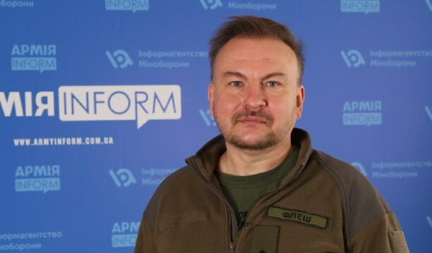 Чоловік у військовому одязі позує на фоні інформаційного стенду «АРМІЯ INFORM». Його зовнішній вигляд підкреслює офіційний характер інтерв’ю або презентації.