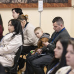 Учасники заходу сидять у залі, уважно слухаючи виступи, серед них присутня й дитина. Делегати мають бейджі, що підкреслює офіційний характер події.