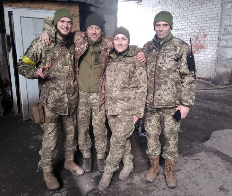Військовослужбовиця разом із побратимами перебуває у польових або фронтових умовах, зберігаючи бойовий дух навіть у складному середовищі. Спільне фото підкреслює товариськість, підтримку та єдність військового колективу.