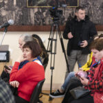Частина аудиторії слухає виступи, деякі учасники користуються телефонами або роблять нотатки. У приміщенні також працює відеооператор, який фіксує подію.